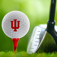 Golf ball with Indiana University trident sitting on a golf tee with golf club next to the ball and tee.