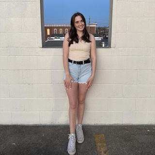 Senior Hannah Scott is wearing a cream colored top and blue denim shorts and is standing in front of a window against a wall.